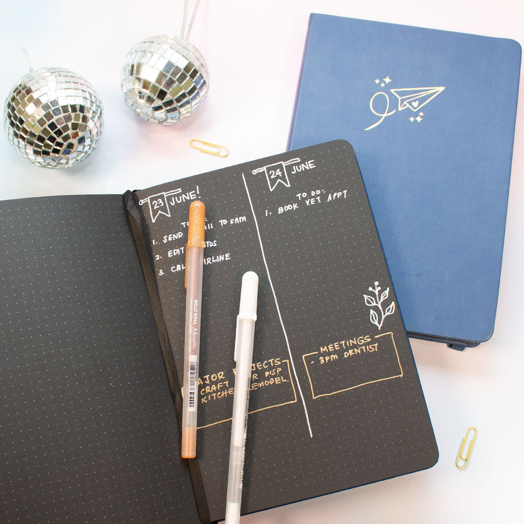 black paper dot grid journal with writing with two gel pens on top two small disco balls above the journal and a closed black paper dot grid journal with a paper plane design on front