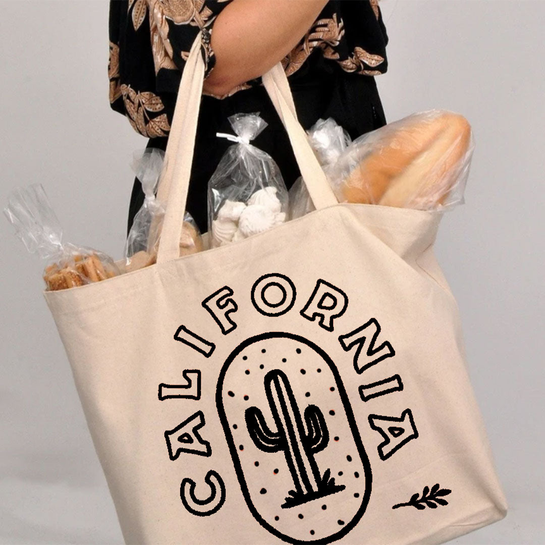 someone holding the white cream colored tote bag with a black cactus print and the word California in black writing wrapped around the cactus with bread and other goodies poking out of the top of the bag