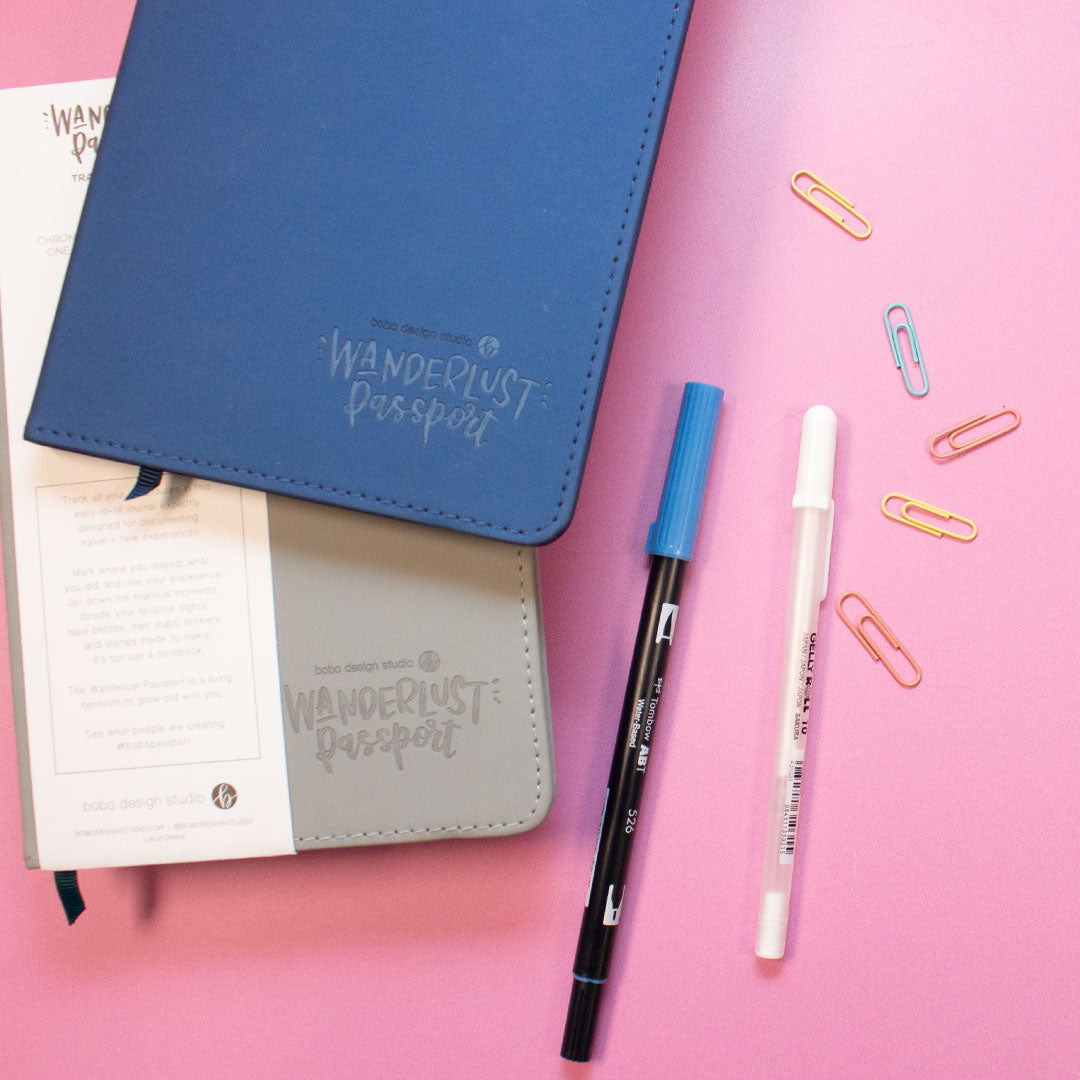 a blue wanderlust passport on top of a grey wanderlust passport with two pens next to them and five small paperclips