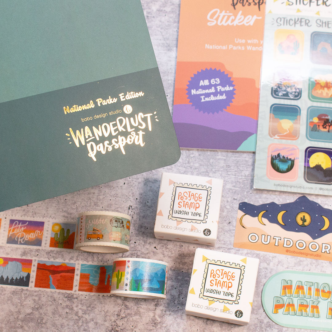top left corner a small corner of the dark green national parks wanderlust passport underneath are two postage stamp washi tapes rolled out with their boxes they come in right next to them. next to the wahi tape boxes are two stickers semi out of frame one says outdoorsy and the other says national park nerd. above the stickers are the national park sticker book and sticker sheet