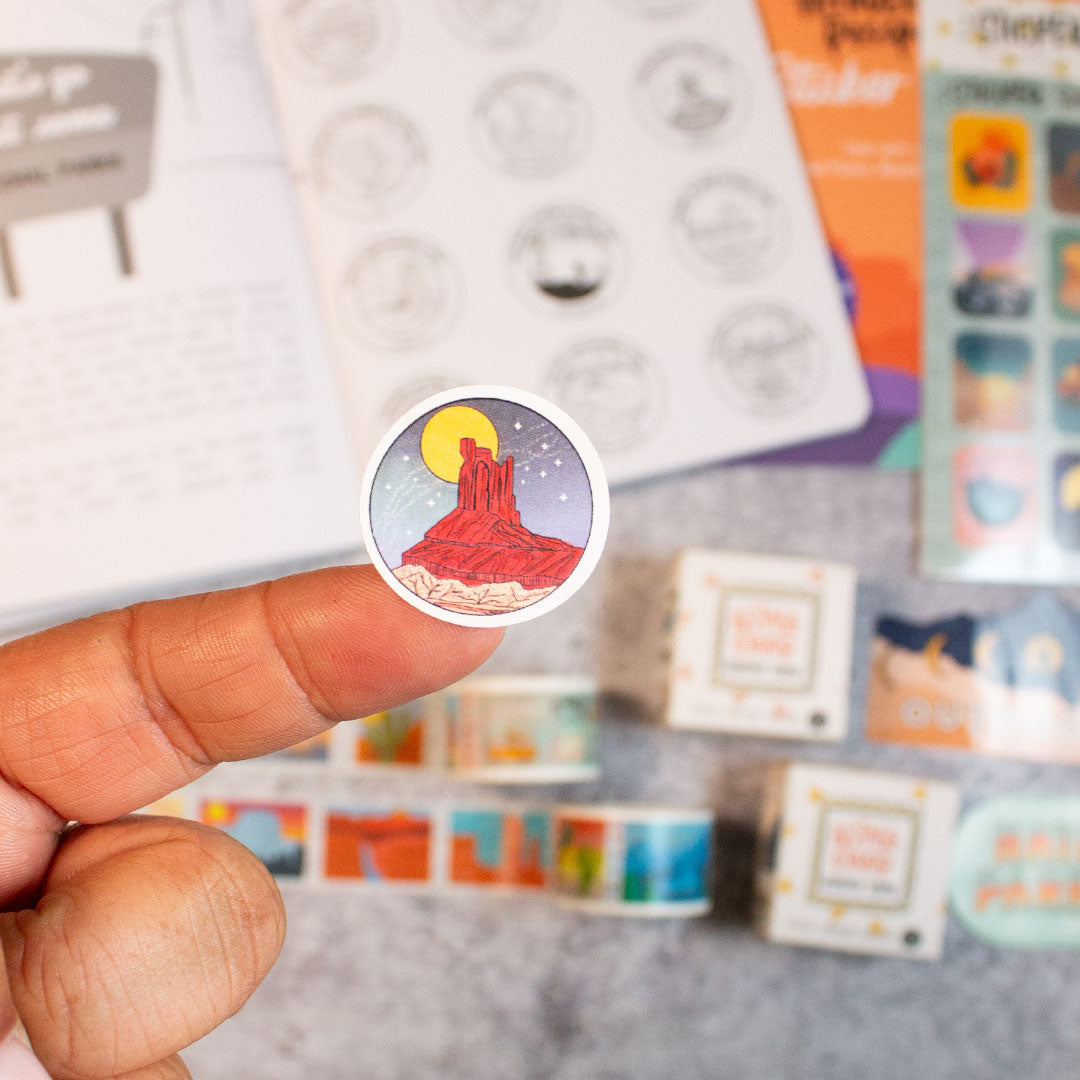 a left finger holding a small circle sticker with a rock mountain. on the table underneath the finger is an open national parks wanderlust passport next to the national parks sticker book and sticker sheet. underneath the passport are two postage stamp washi tape boxes with each washi rolled out. next to the washi tape ypu can see a small amount of the outdoorsy sticker and national park nerd sticker