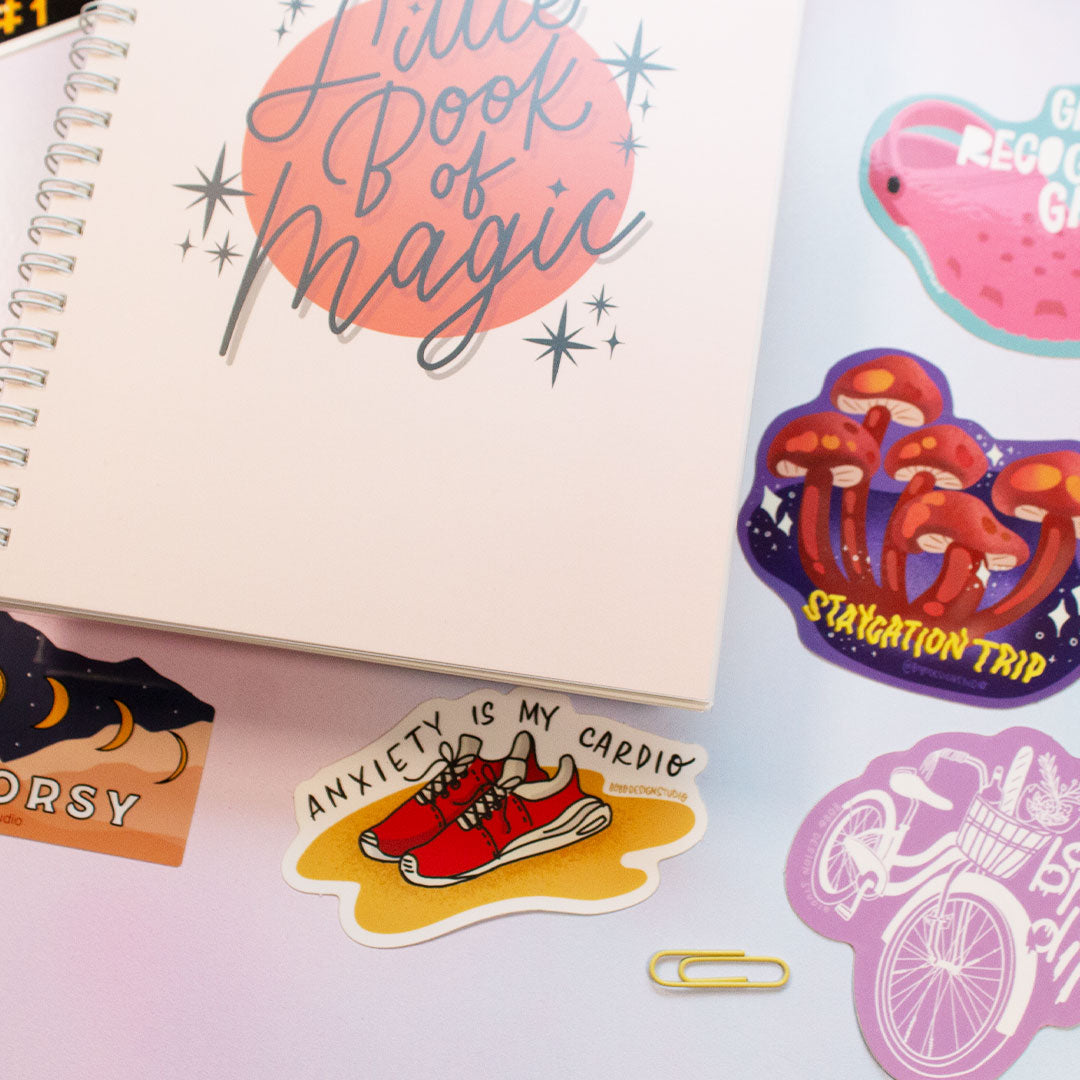 zoomed in picture of reusable sticker book that says "little book of magic" on the front and three stickers to the right of the book and two stickers underneath the book and a small paperclip