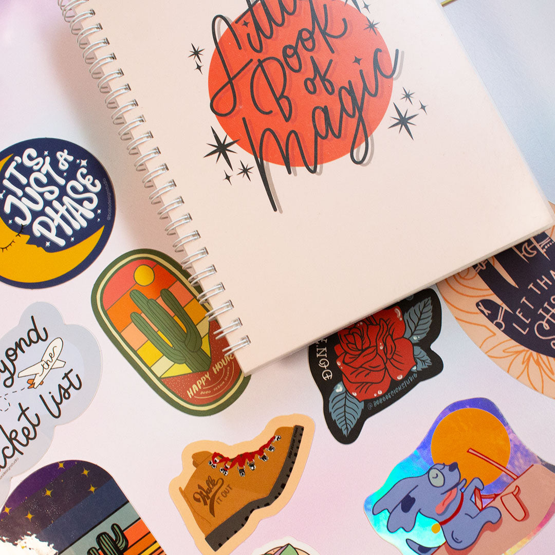 zoomed in image of reusable sticker book that says "little book of magic" on the front and eight stickers in total spread out to the left and underneath the book
