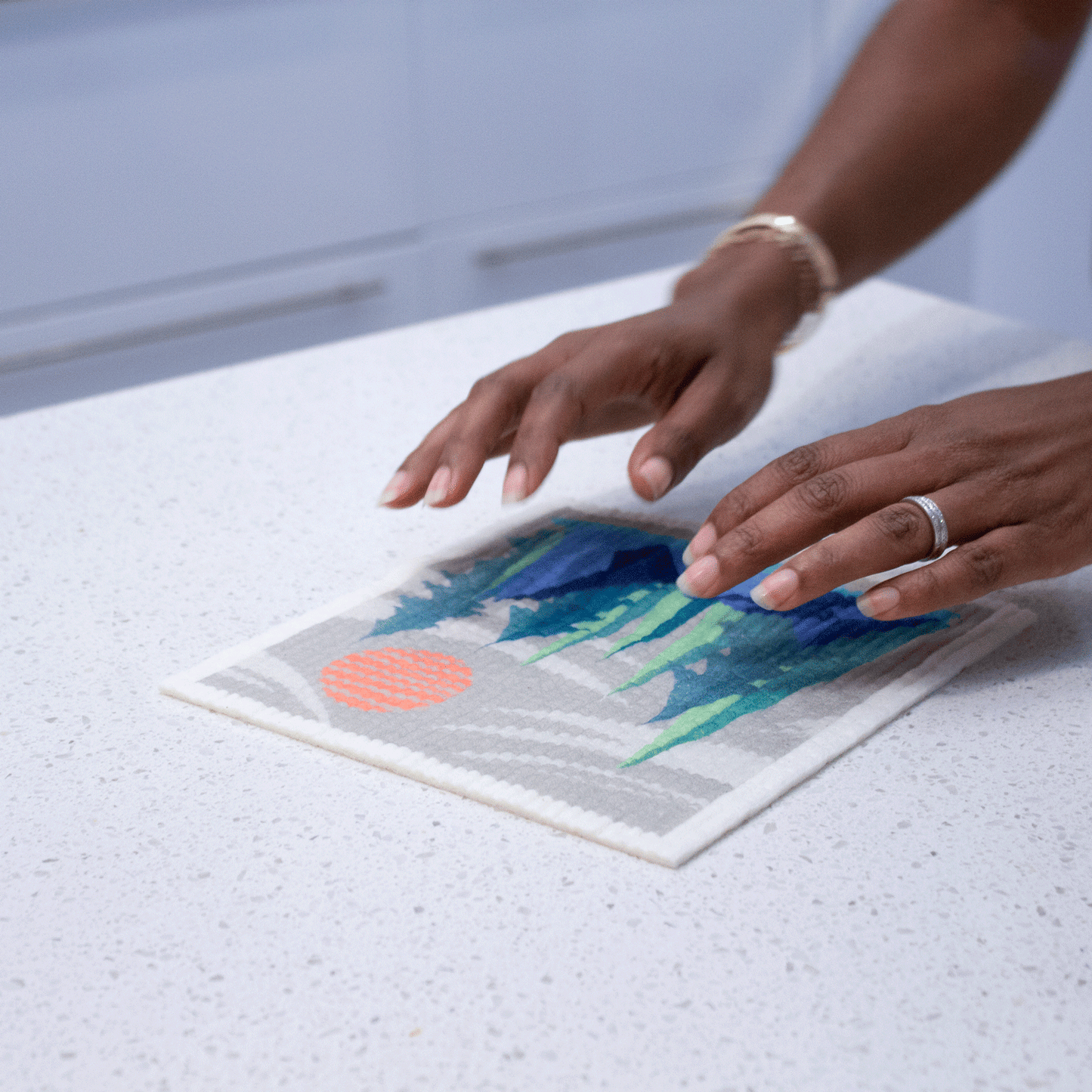 Two hands with gold and silver jewelry hover above the Pacific Northwest sponge cloth. The sponge cloth is on a kitchen counter.