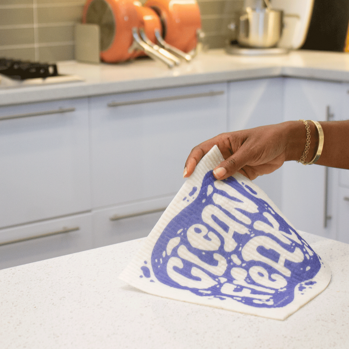 A hand holds a Super Cloth with the Clean Freak design in white and blue over a kitchen counter. The hand is demonstrating how flexible the cloth is when wet. 