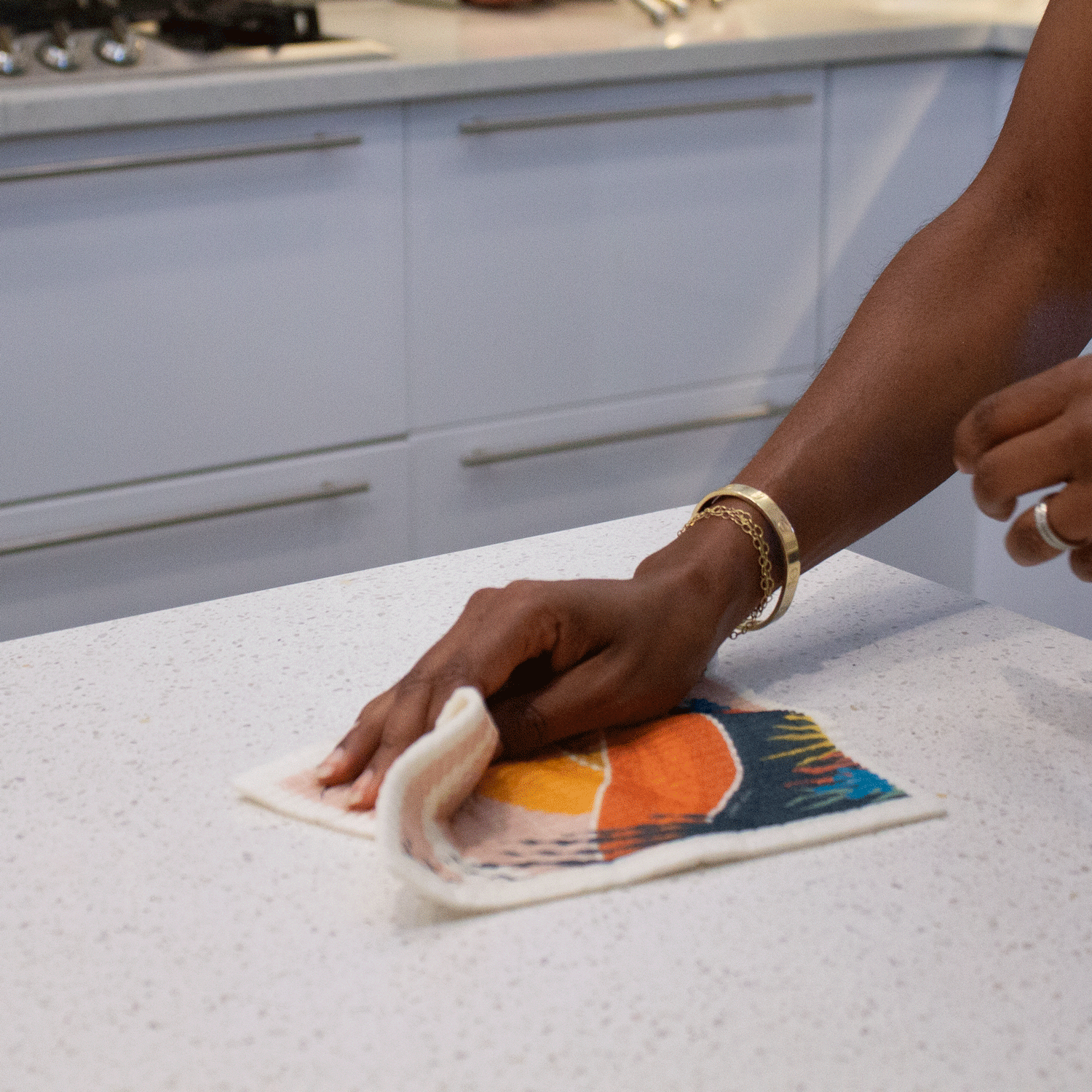 A Hand wipes a kitchen counter with the Desert Oasis sponge cloth. You can see the sponge cloth is flexible as it folds in soft curves. The bright desert scene is partially visible under the hand. 
