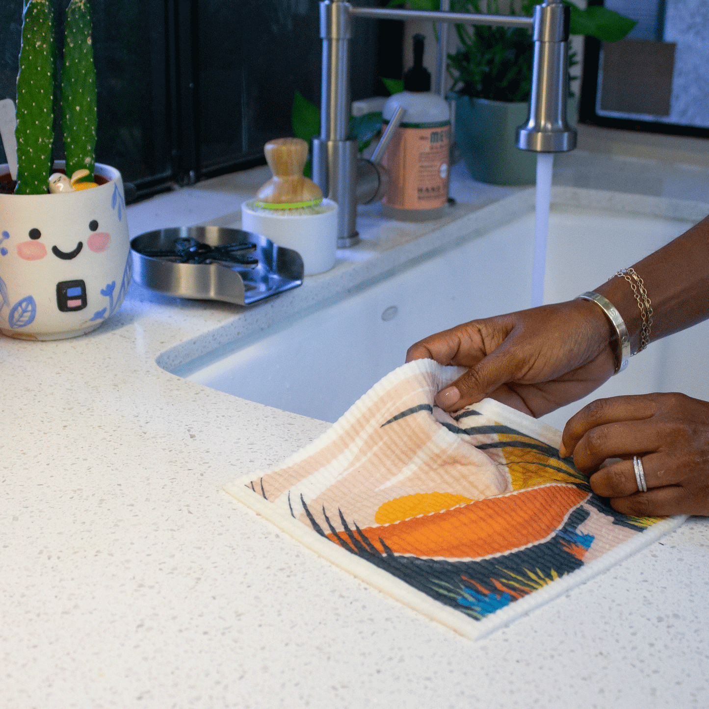 Two hands hold a Desert Oasis Super Cloth near the edge of a sink. A desert scene with hills, plants, and a sun is printed on the sponge cloth. The sponge cloth is bending in soft folds. Kitchen items such as soap, some succulents, and a scrub brush are in the background.