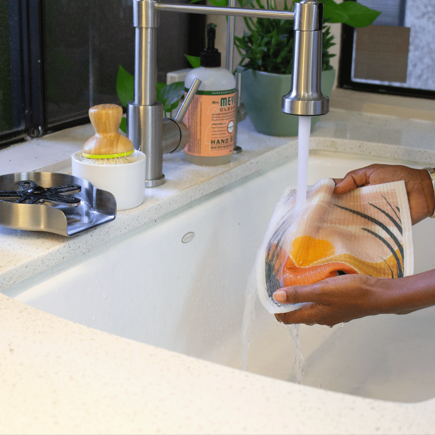 Two hands hold a Desert Oasis super cloth under a kitchen faucet. The water is falling onto the sponge cloth, and you can see that the sponge cloth is bending in soft folds. The bright desert scene, of a sun, hills, and plants; printed on the cloth is visible. There are plants, soap, and other kitchen items in the background.