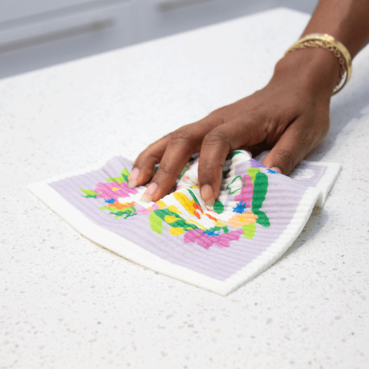 Hand holds “Fresh & Clean” sponge cloth and wipes table. Sponge cloth is wet and bunched in the hand’s grasp. The lavender background and flower motif is visible, but the wording is obscured by the hand. 