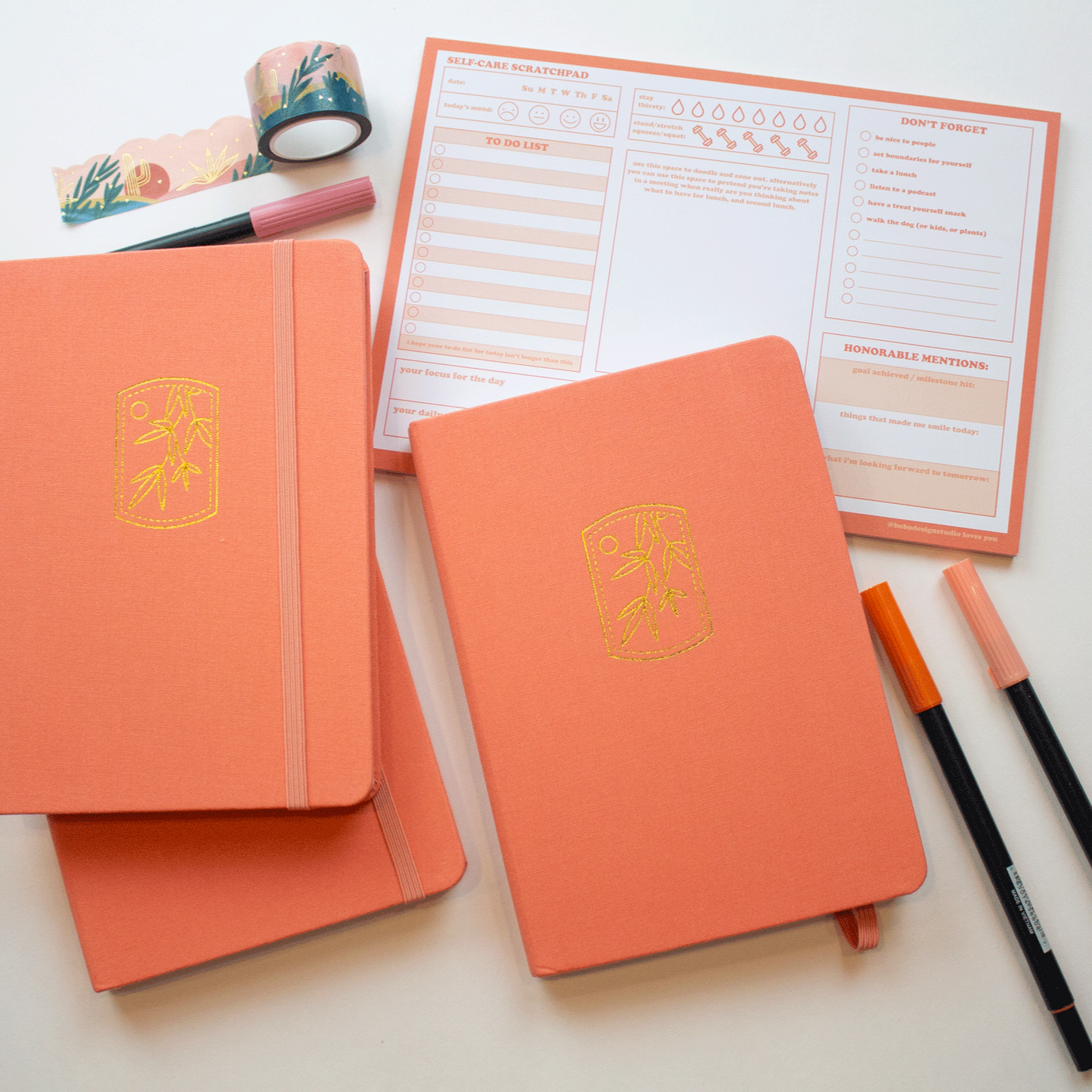 Three tangerine bobo BuJo dot grid journals are stacked near other orange desk supplies. Two books have their elastic closure closed, one book has it pulled open.