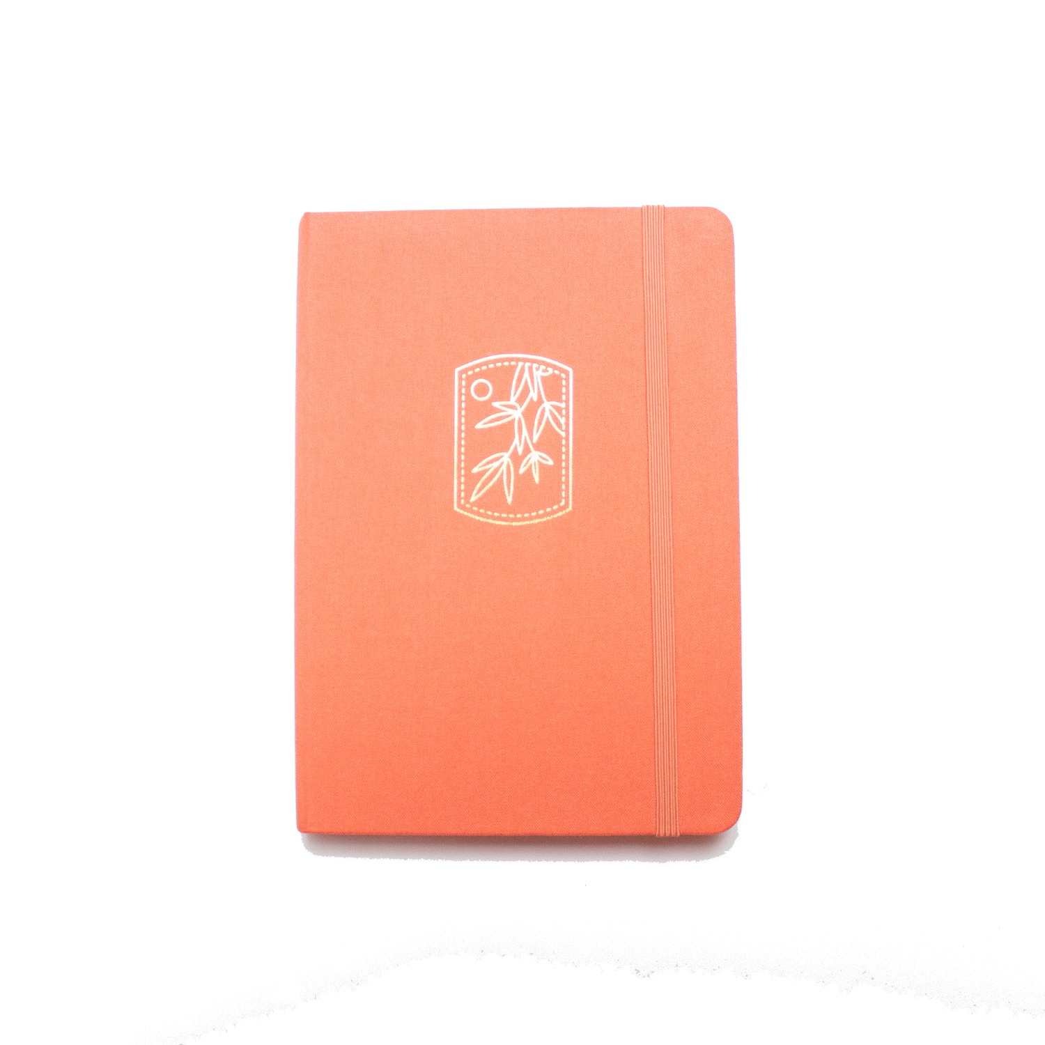 The A5 bobo BuJo Dot Grid Journal in "Tangerine" orange. A gold foil illustration of mandarin tree leaves and a sun is stamped on the front cover. A matching orange elastic closure is holding the journal closed.