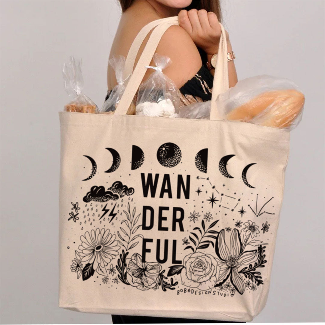 someone holding large canvas tote white cream color with black decals such as the moon cycle, flowers, stars, and clouds with the word "wanderful" spelled out with bread and other various goodies sticking out of the top