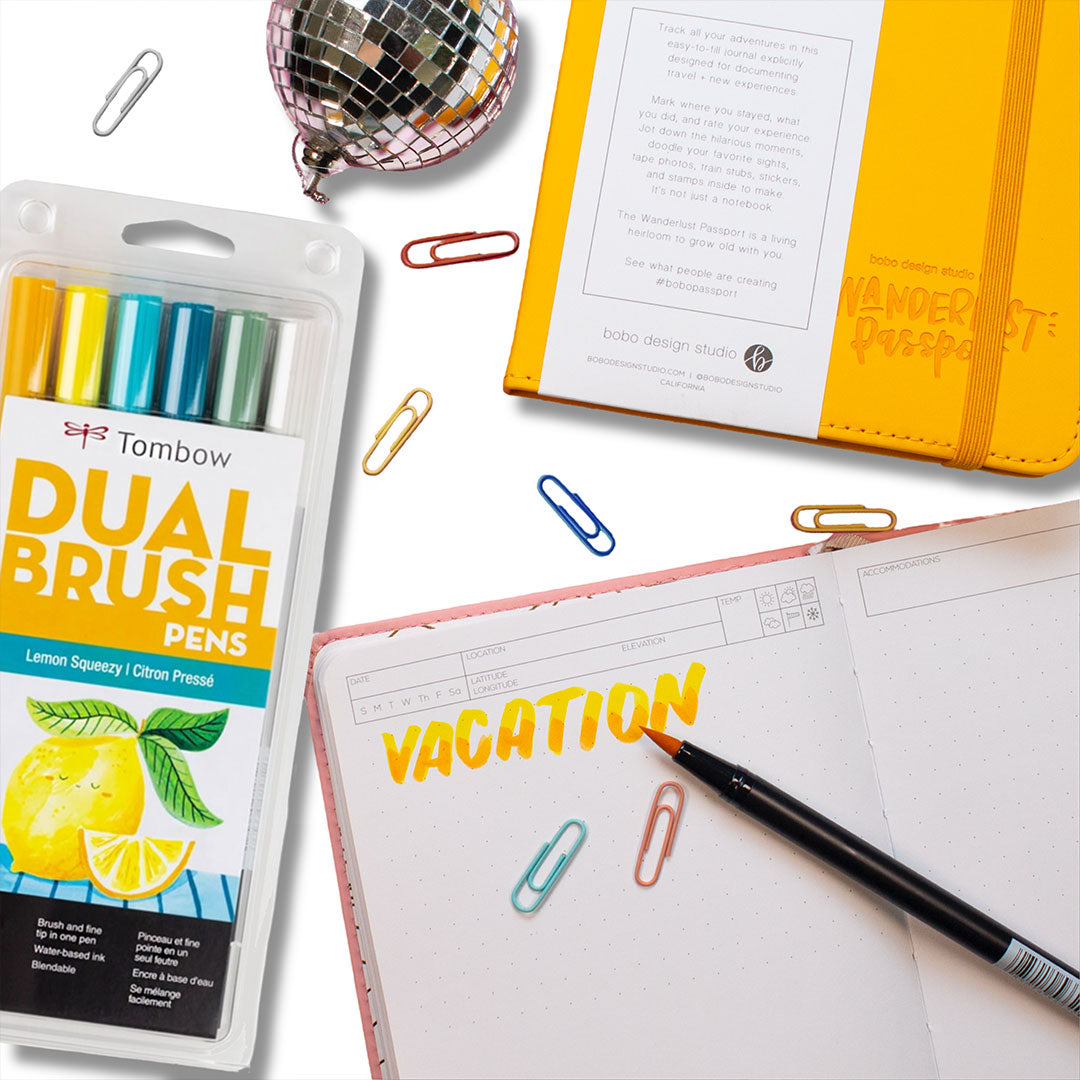 tombow dual drush pens pack with a mini disco ball above and seven small colorful paper clips spread out all over. a small corner of a yellow wanderlust passport showing on the top right and an open pink wanderlust passport with "vacation" written on a page in yellow with an open brush pen on the page