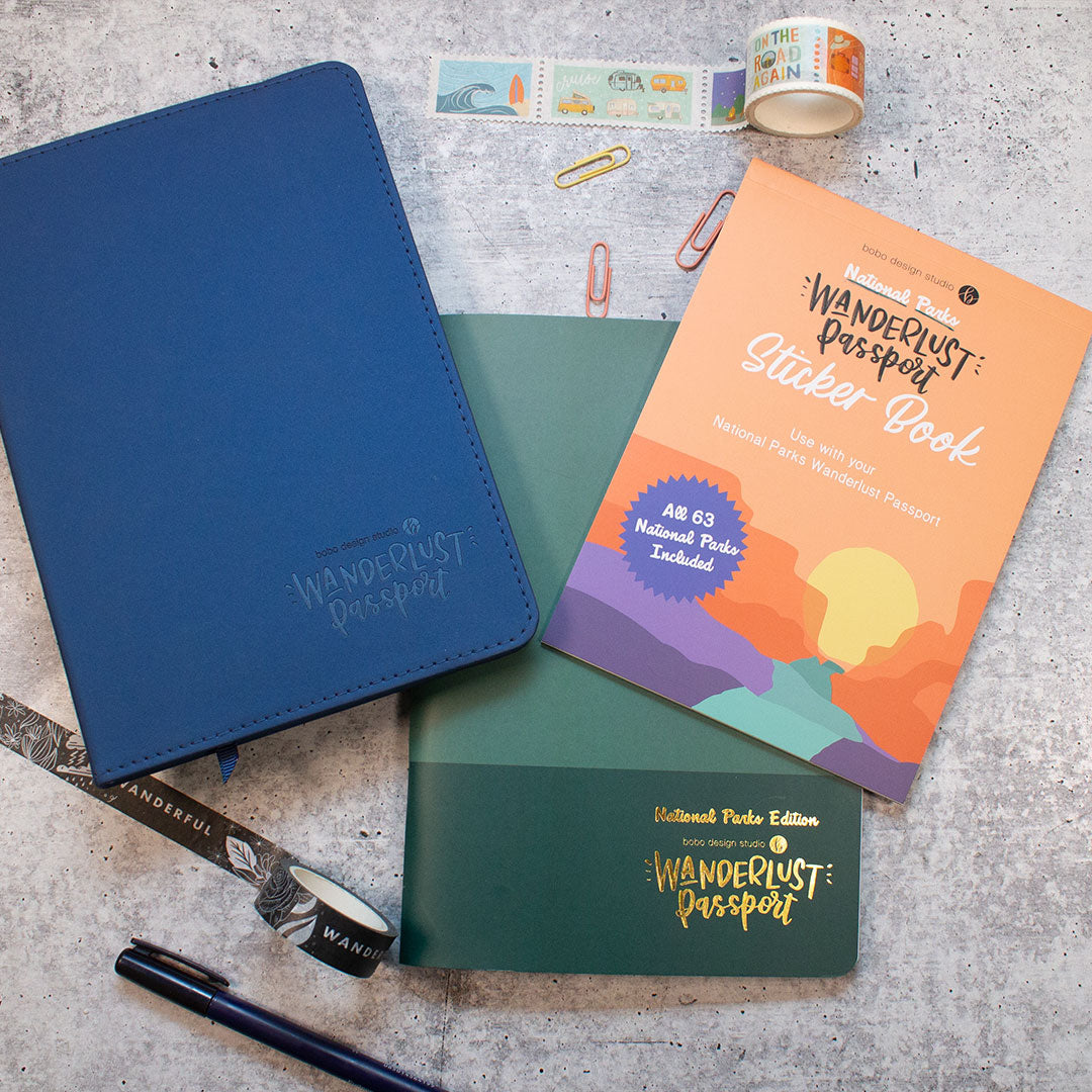 a blue wanderlust passport and a national parks sticker book on top of a national parks wanderlust passport with a rolled out washi tape and three paper clips above and a rolled out washi tape and pen below