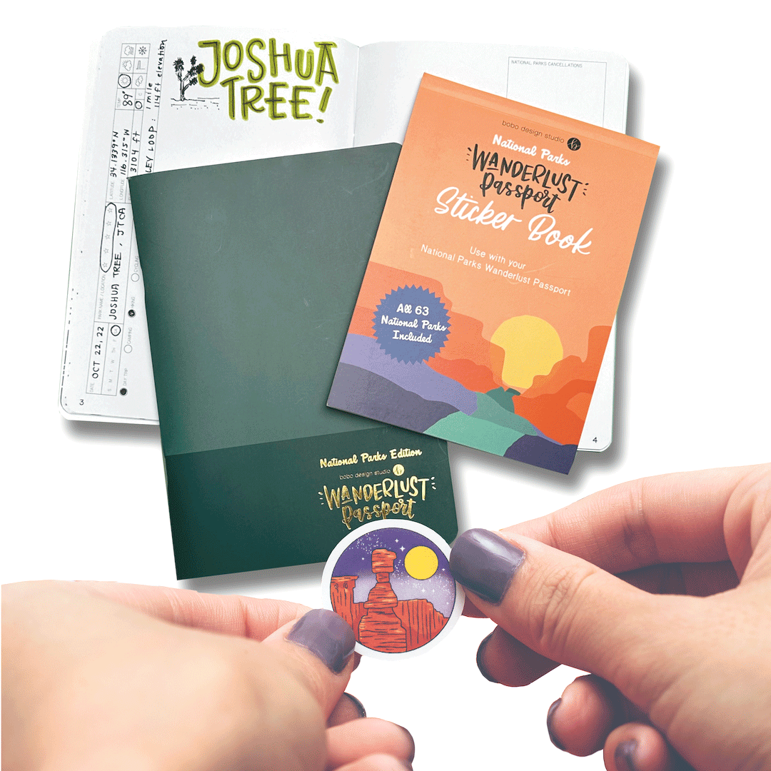 two hands holding a small circle sticker of a rock with the moon behind it. on the table below is the national parks sticker book on top of a closed national parks wanderlust passport on top of a open national parks wanderlust passport that says joshua tree on the top