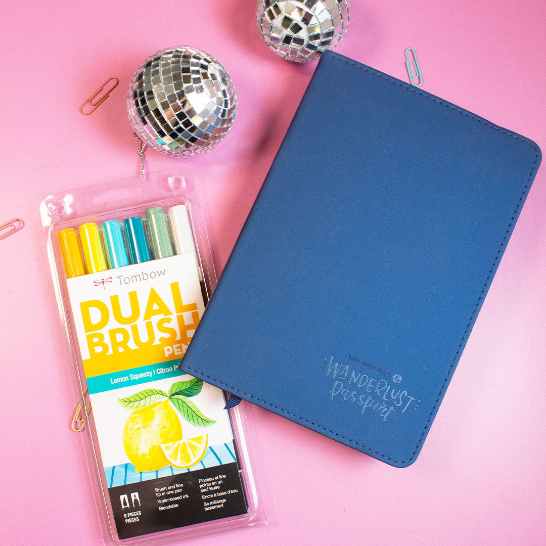 blue wanderlust passport closed with two mini disco balls next to it and tombow duel brush pens set slightly underneath it and four small paper clips sprinkled in the background