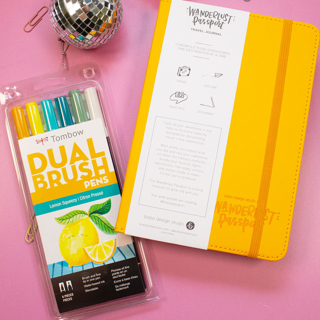 yellow wanderlust passport closed with a mini disco ball next to it and a tombow dual brush pen set slightly underneath the passport with two mini paper clips in the background