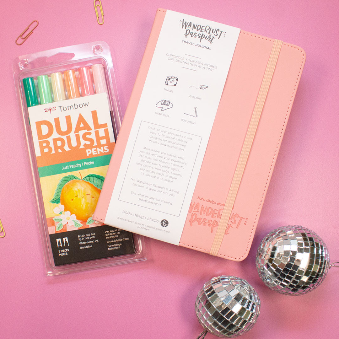 pink wanderlust passport closed on top of a tombow dual brush pen pack and two small paper clips above the pens. two mini disco balls underneath the passport