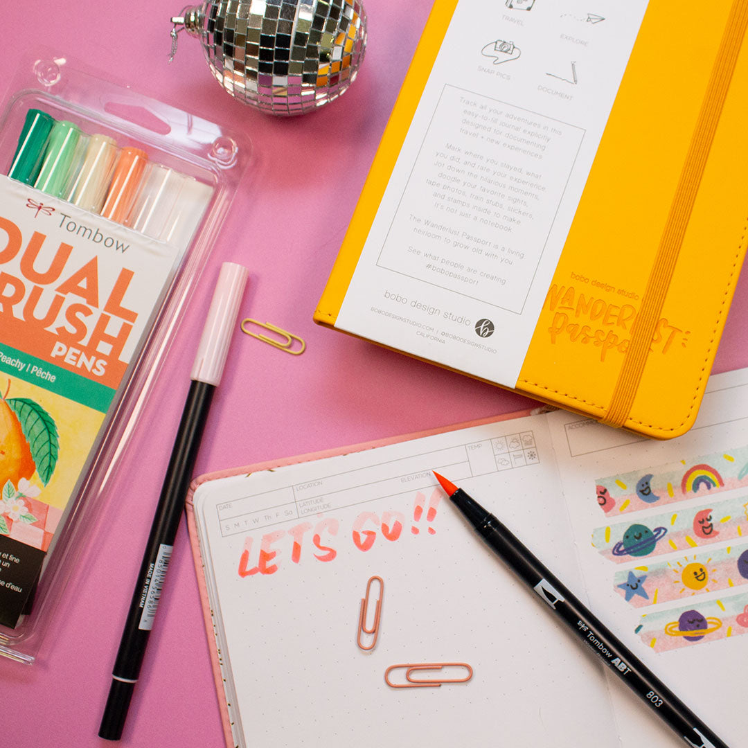 yellow wanderlust passport with a mini disco ball next to it and tombow dual brush pen pack with one pen next to the pack. underneath the passport is an open pink wanderlust passport that says lets go written in pink with three mini paper clips spread out. on the other page is friendly skies washi tape rolled out