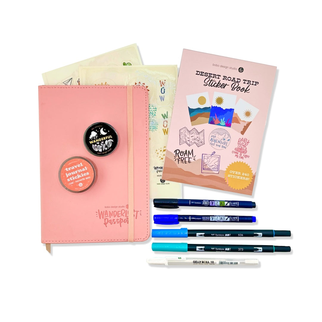 a pink wanderlust passport on top of two sticker sheets and next to a desert road trip sticker book and two blue tombow duel brush pens, a black and a blue tombow fudenosuke brush pen, and a white gelly roll gel pen. on top of the passport are a black wanderful washi tape and a clear travel journal washi tape