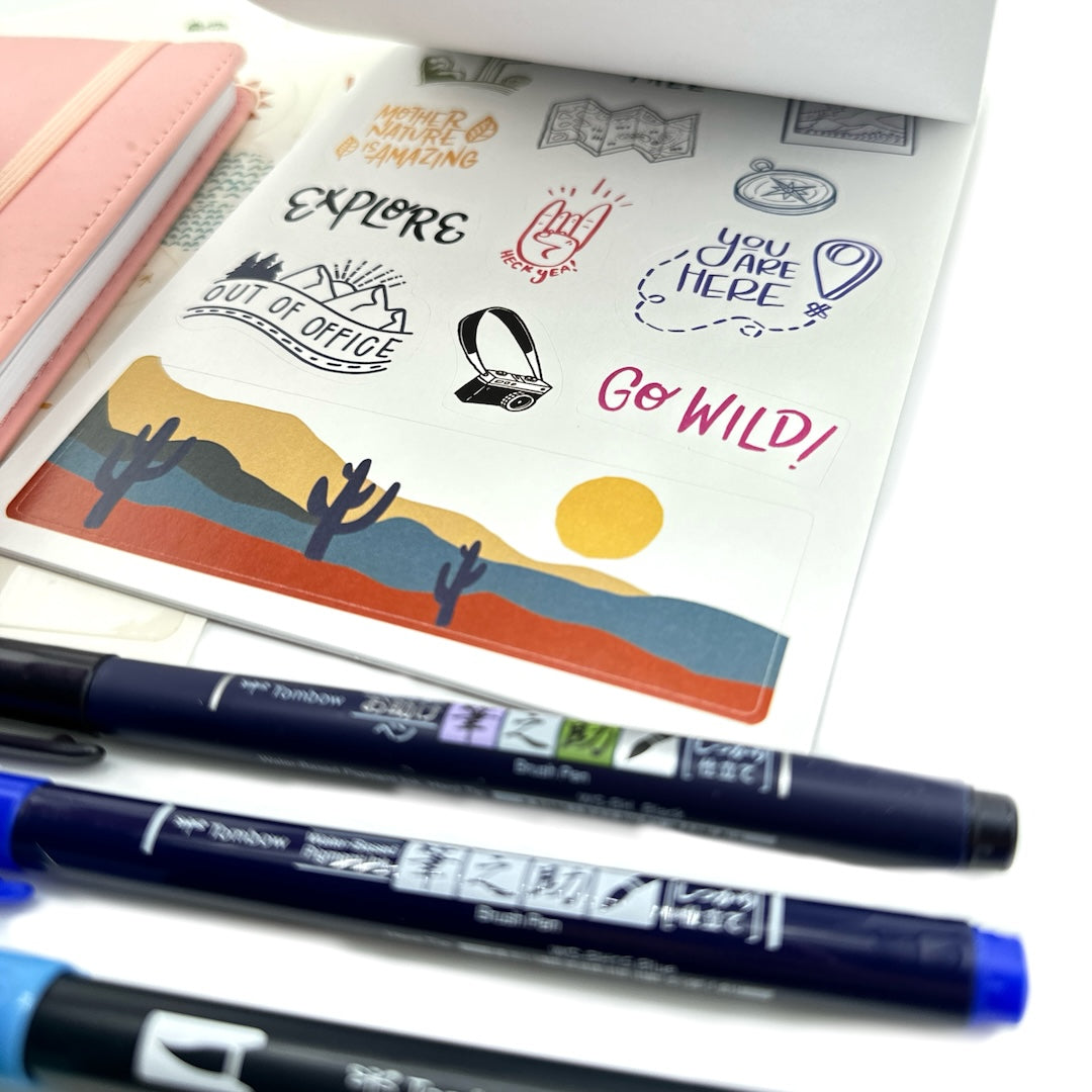 an open desert sticker book next to a pink wanderlust passport with two blue pens and one black one underneath the sticker book