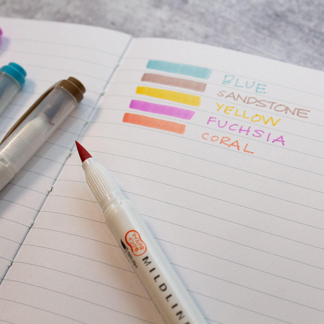 open lined journal with mildliner brush pens swatched with the names of the shades next to it and an open pen on the page with two closed ones on the other page