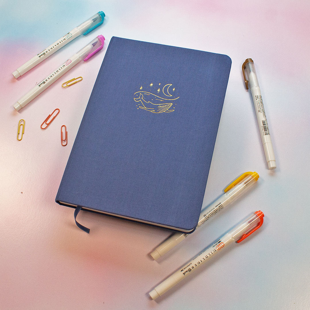 blue journal with a gold whale decal on the front with a moon two pens and four paperclips to the left and three pens to the right