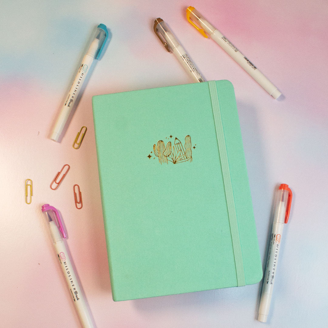 teal journal with a pencil and cactus decal in gold on the front on the left are two pens and four paper clips, two pens above the journal and one pen on the bottom right