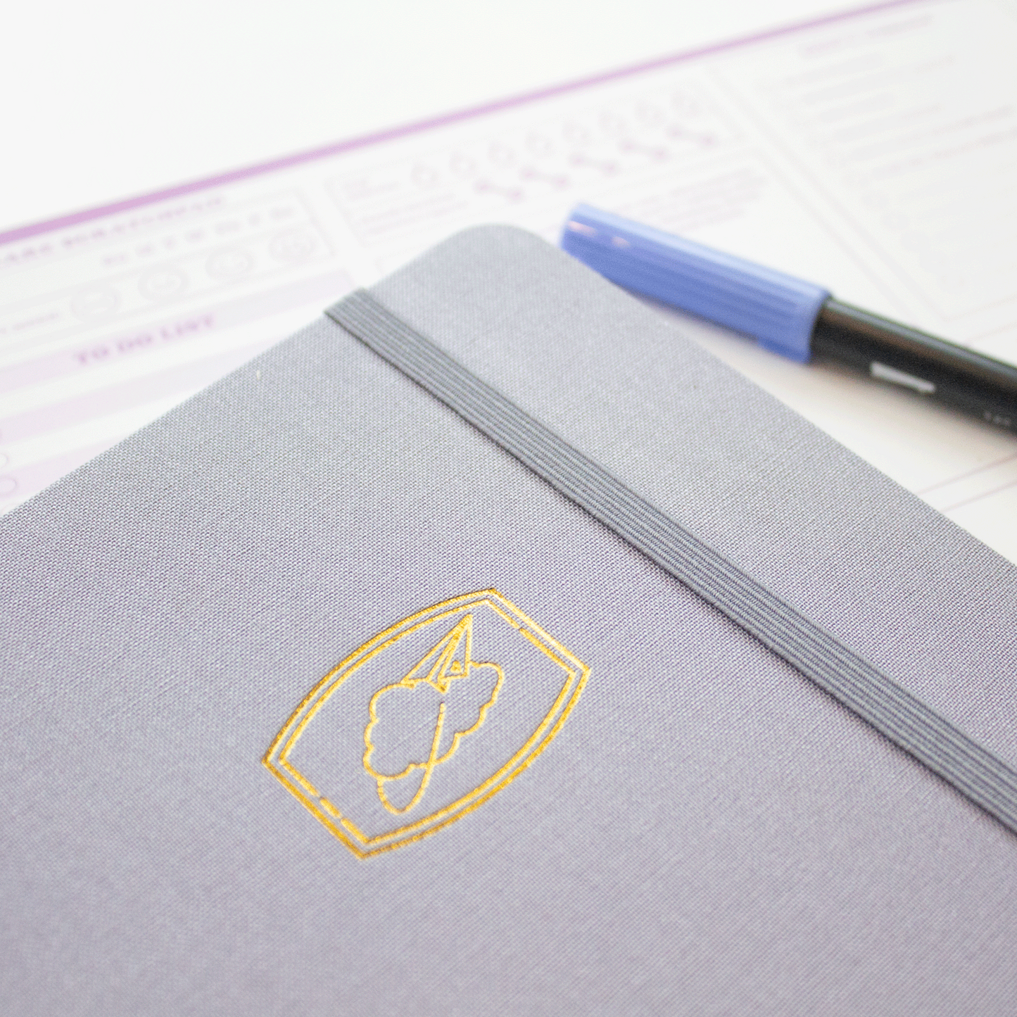 A close up of the front cover of the Cloud 9 light blue bobo BuJo dot grid Journal. The corresponding blue elastic closure is holding the journal closed. The gold stamped cloud illustration is on the front cover. A marker and bobo self-care scratch pad are in the background.