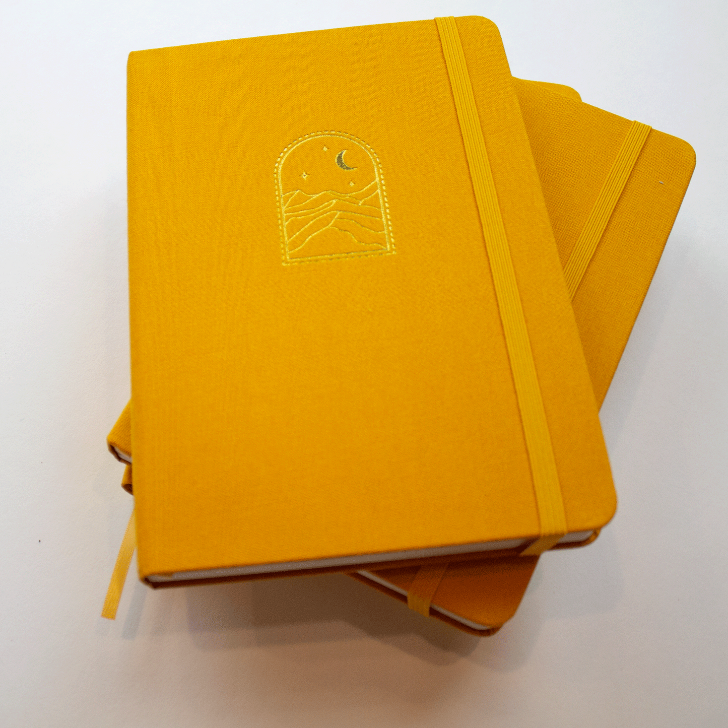 A stack of three mustard yellow "Golden Dunes" bobo dot grid journals. The gold stamped starry night sky and sand dune illustration on the front cover can be seen on the first book. Their matching yellow elastic closures and ribbon bookmarks can also be seen.