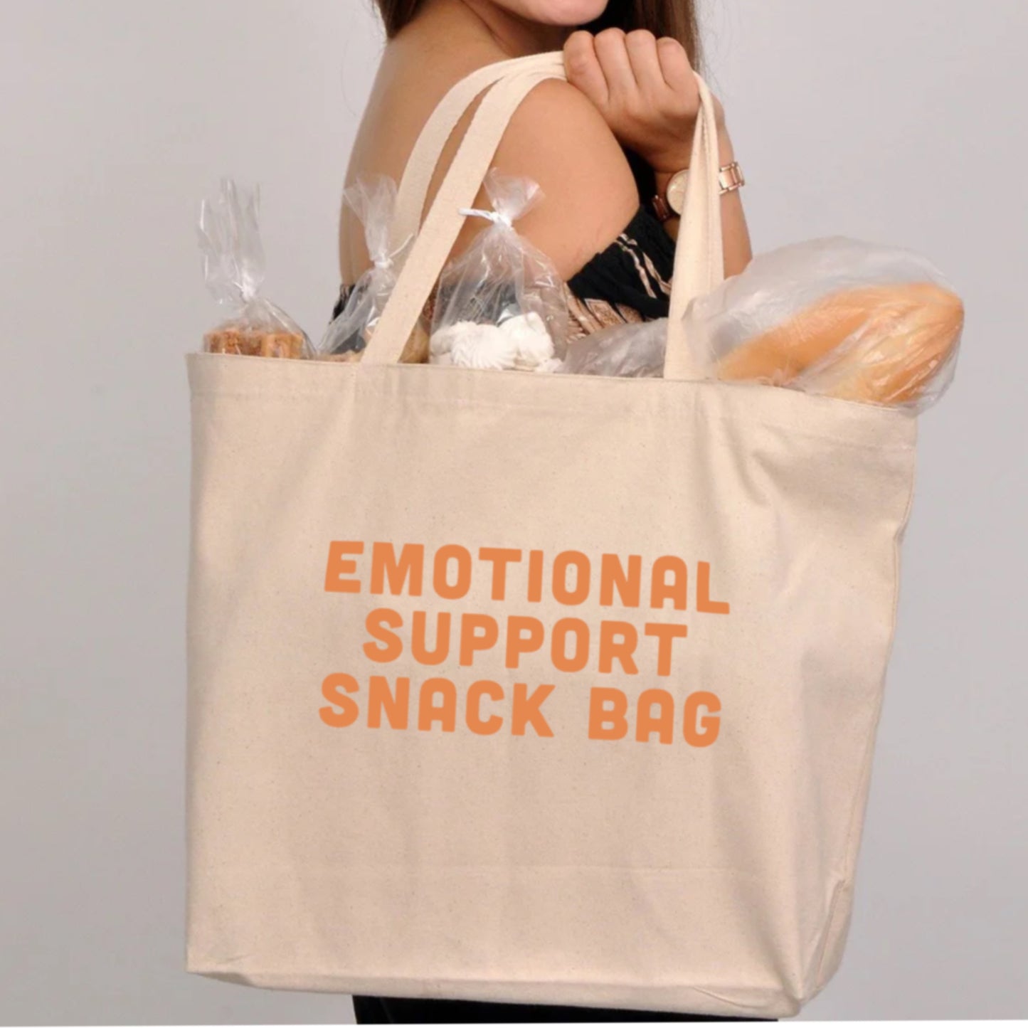 a person holding white cream canvas tote bag with the words "emotional support snack bag" in orange lettering with bread and other goodies peeking out of the top of the bag
