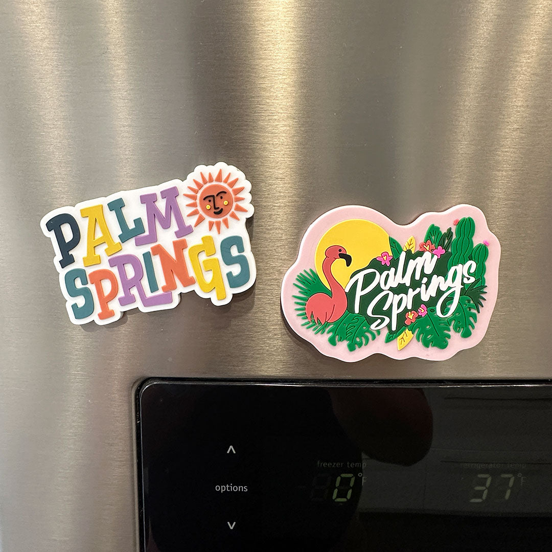 Palm Springs flamingo magnet. features a flamingo with a sun behind it and leaves with some cactus and "palm springs" written in white cursive letters with a pink background on a fridge along with a white magnet that says "palm springs" in multiple colors and a orange sun with a smiling face beside it