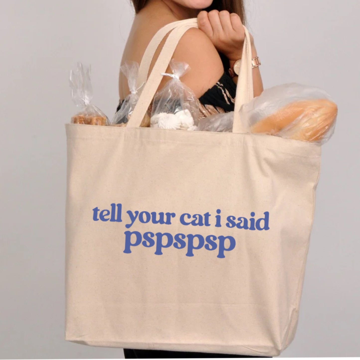someone holding cream white canvas tote bag with " tell your cat i said pspspspsp" in blue lettering with bread and other goodies sticking out of the top of bag