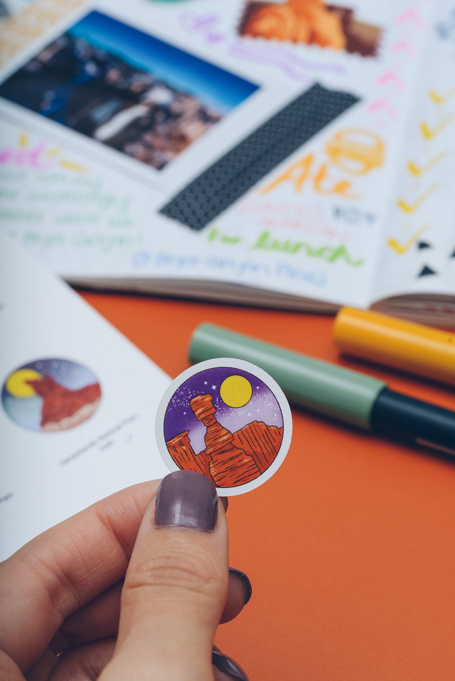 a left hand holding a small circle sticker with a rock mountain and a moon. in the background a small corner of the national parks wanderlust passport sticker book and an open filled in journal and two tombow pens