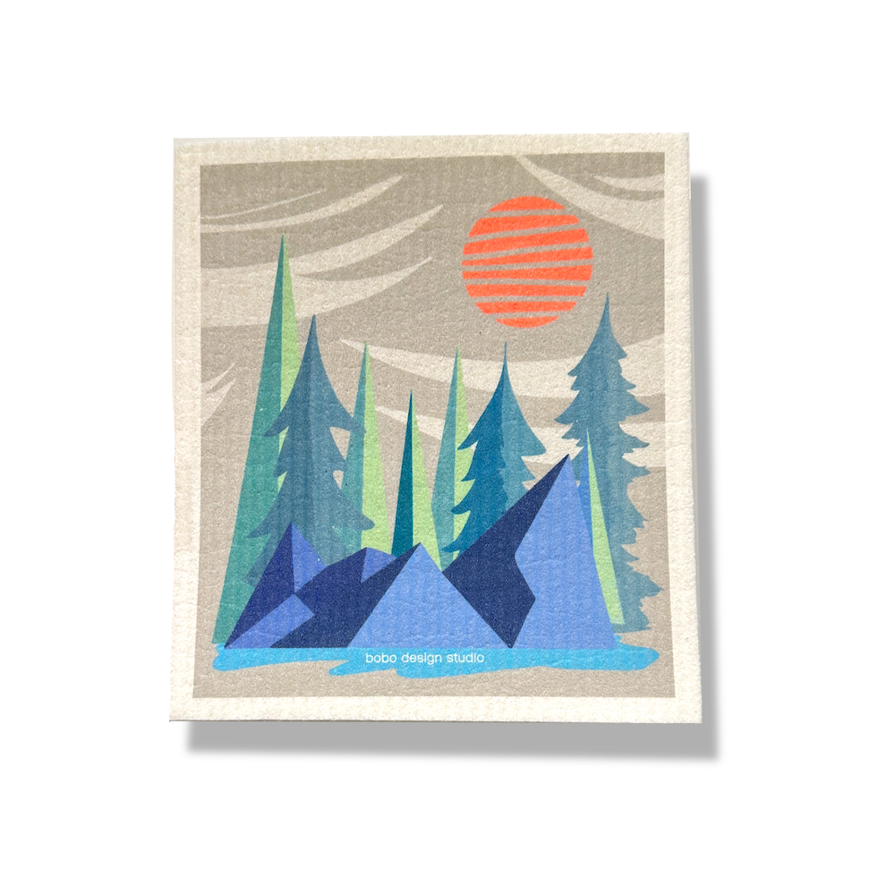 Portrait orientation sponge cloth (6.5x8 inches) with vertical wrinkled spongy texture. Printed image centered on cloth with white border. Image of grey sky with abstract white clouds, orange sun, green trees. Dark blue geometric rocks and an aqua blue pond in the foreground.