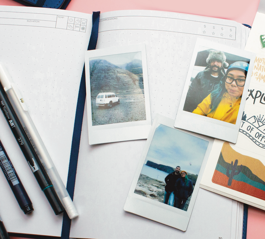 a blank open navy blue wanderlust passport with three polariod pictures on top, three pens as well as as sticker sheet