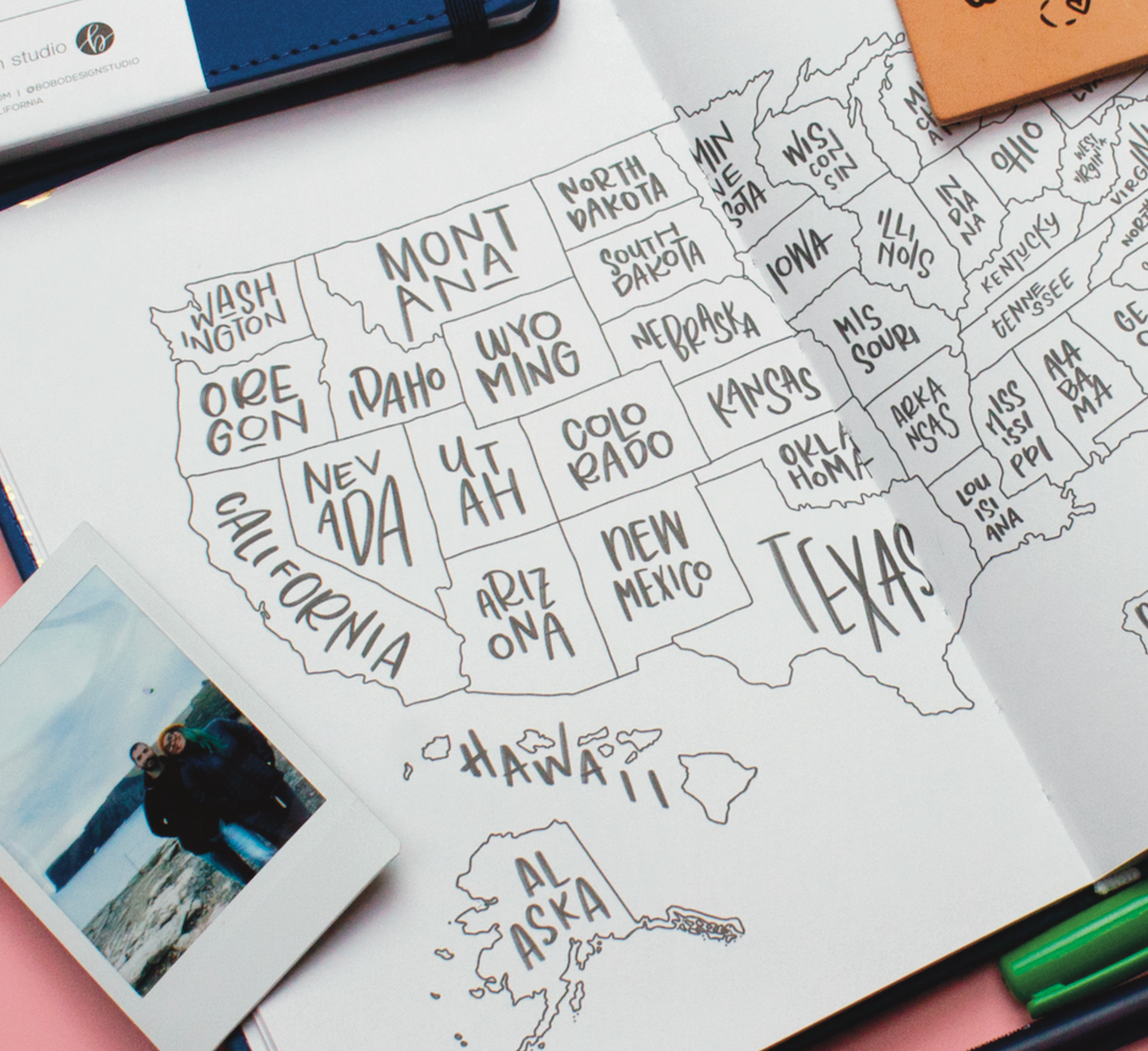 a blue open wanderlust passport showing a map of the united states with a small polaroid picture on top of it, a closed passport showing slightly on top with two pens on the bottom