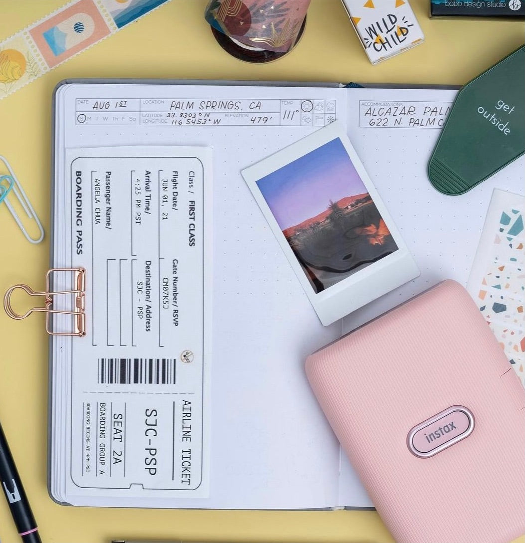 Wanderlust Passport Travel Journal open to a journal page in progress, the page's location, weather, and date tracker if filled-in, and the page is being decorated with washi tape, a small printed photograph of a mountain and trees, a boarding pass, and washi stickers. Markers, paperclips, a photo printer, and a "get outside" keychain are around it.