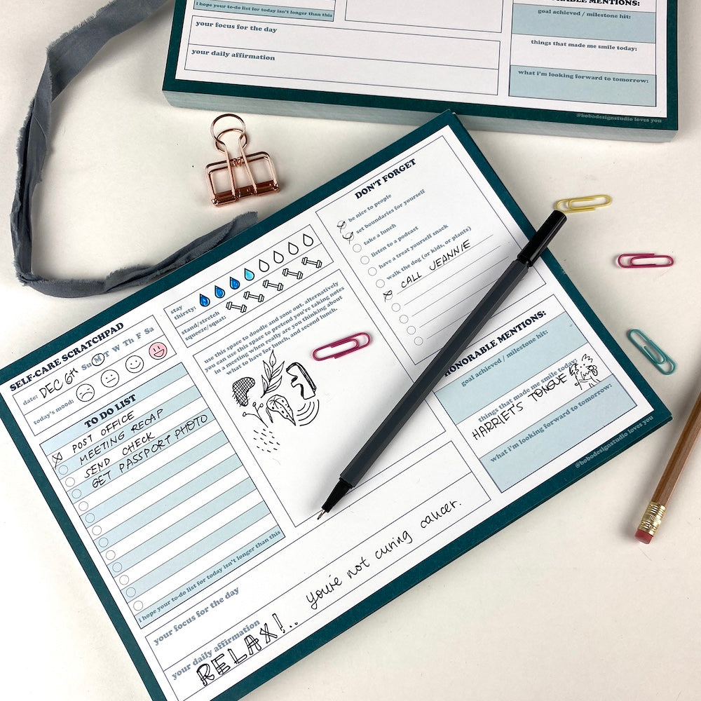 2 Teal Scratch Pads on a desk with paperclips, a pen, a pencil, and a binder clip nearby. Some of the three columns of self-care daily planning trackers are filled in. The daily mood emoji is colored in. Some of the waterdrop hydration tracker is filled in. The to-do list section is filled in with a handwritten example list Post Office, Meeting Recap, Send Check, Get Passport Photo. 