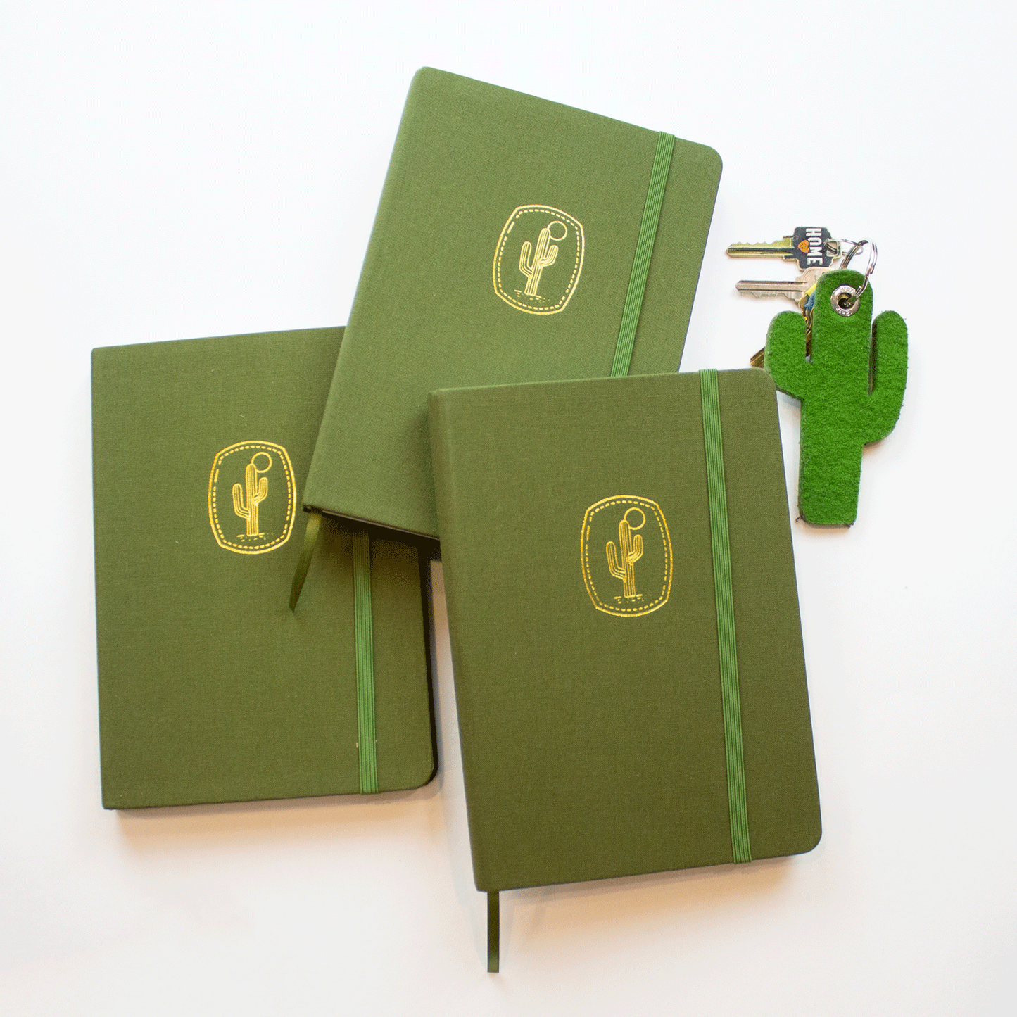 Three bobo BuJo dot grid Journals in Cactus green. The journals are piled together in a row, and their ribbon bookmarks and elastic closures are visible. A green saguaro cactus keychain with keys sits to their right.