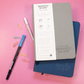 a grey wanderlust passport on top of a blue wanderlust passport with two pens next to them and five small paperclips