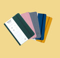 all five of our wanderlust passport colors on top of each other spread out like a fan. from left to right dark green, light pink, light grey, navy blue, and yellow