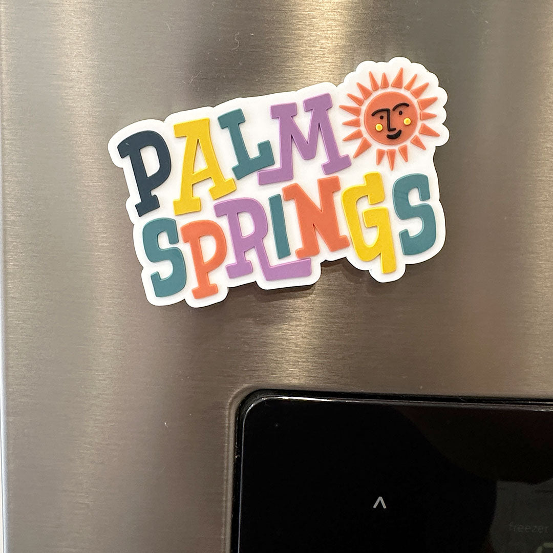 zoomed in image of white magnet that says "palm springs" in different colors for each word and a orange sun with a smiling face on the top right corner next to the word "palm" on a fridge