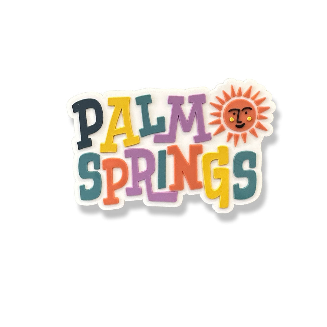 white magnet that says "palm springs" in different colors for each word and a orange sun with a smiling face on the top right corner next to the word "palm"