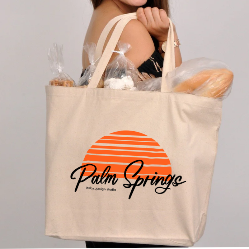somene holding white cream colored canvas tote bag with "palm springs" written in black font cursive with an orange half circle that looks like a top part of a sunset on their shoulder with bread and other baked goods sticking out of the top of the bag