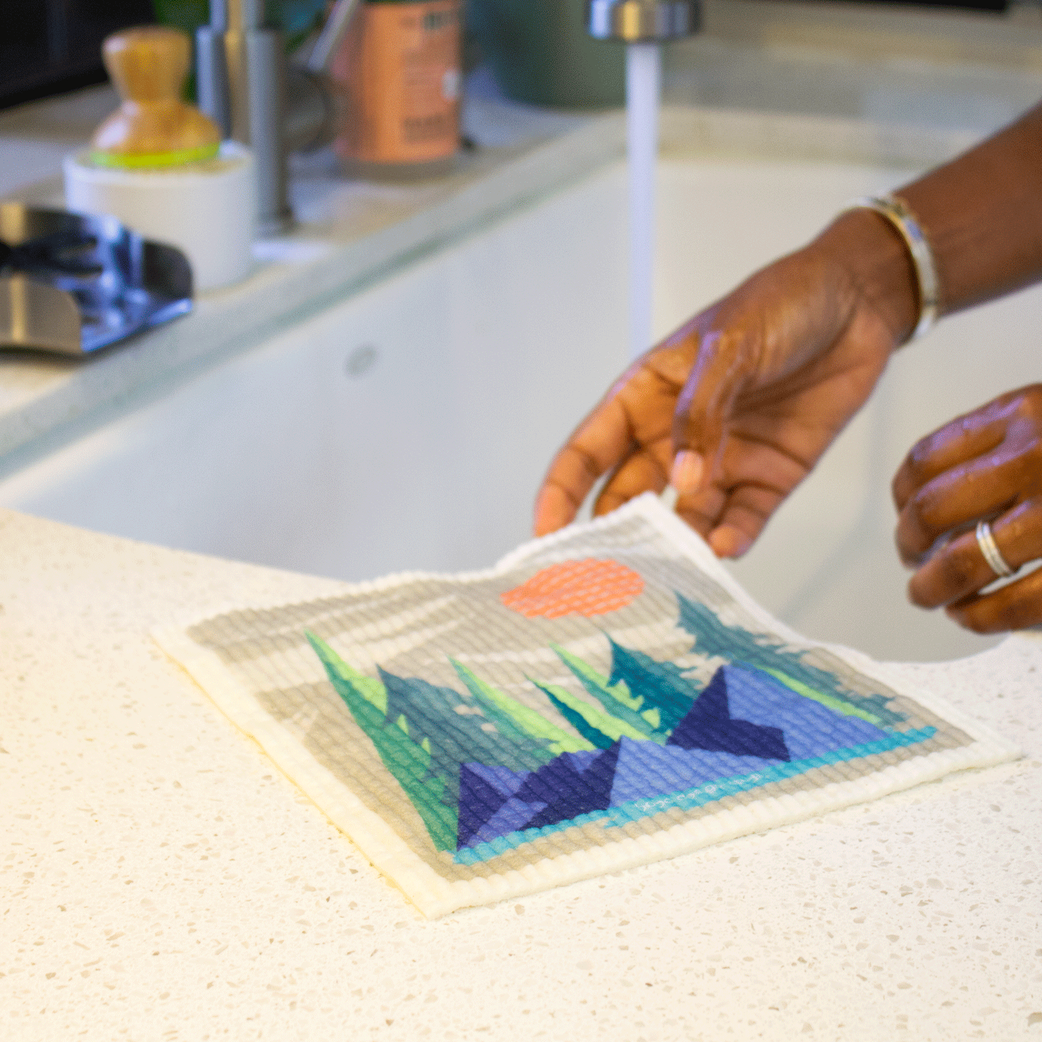 Two hands hold the Pacific Northwest sponge cloth near the edge of a kitchen sink. There is an abstract geometric forest scene printed on the sponge cloth.