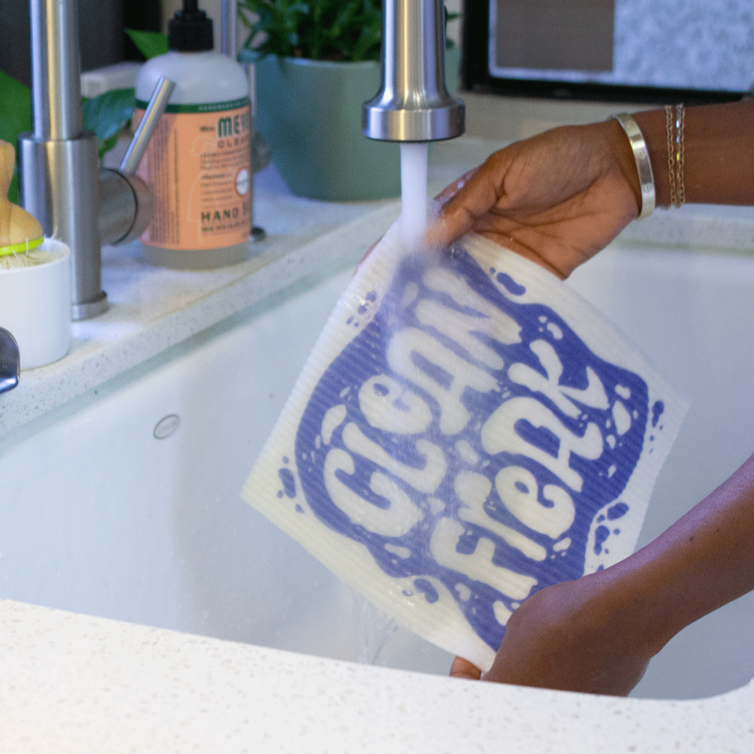 Two hands hold the Clean Freak Super Cloth under a running kitchen faucet. The blue and white clean freak print is partially obscured by the running water.