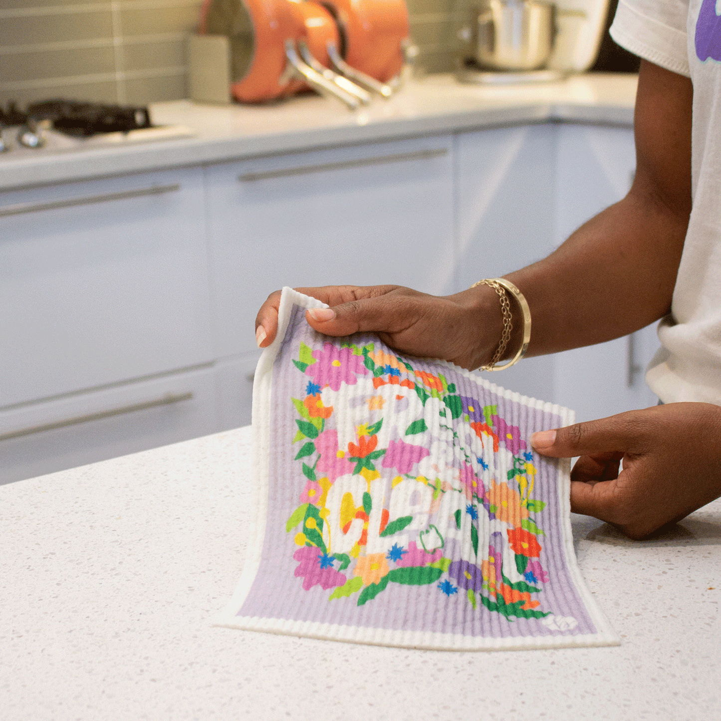 Hands hold sponge cloth in a white cabinet modern kitchen. Sponge cloth says “Fresh & Clean” surrounded by colorful abstract flowers. Sponge cloth is wet and is now soft and draping (no longer dry and flat).