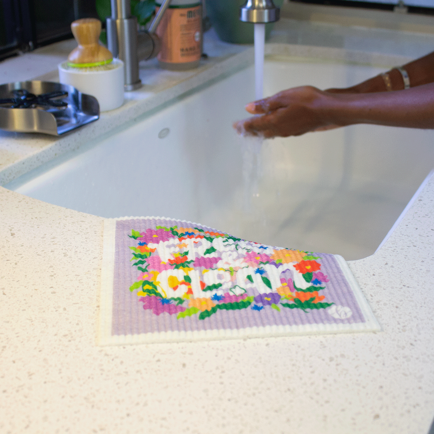 Sponge cloth on the edge of a sink, someone is washing their hands in the background. Sponge cloth has a printed design: “Fresh & Clean” with colorful flowers. Sponge cloth is wet and flexible and the top right corner drapes into the sink.