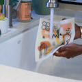 The Mid Mod Super Cloth is being held under a kitchen faucet. Two hands hold the sponge cloth as it folds in soft curves under the running water. You can see the abstract printed design on the cloth. Kitchen items are in the background.